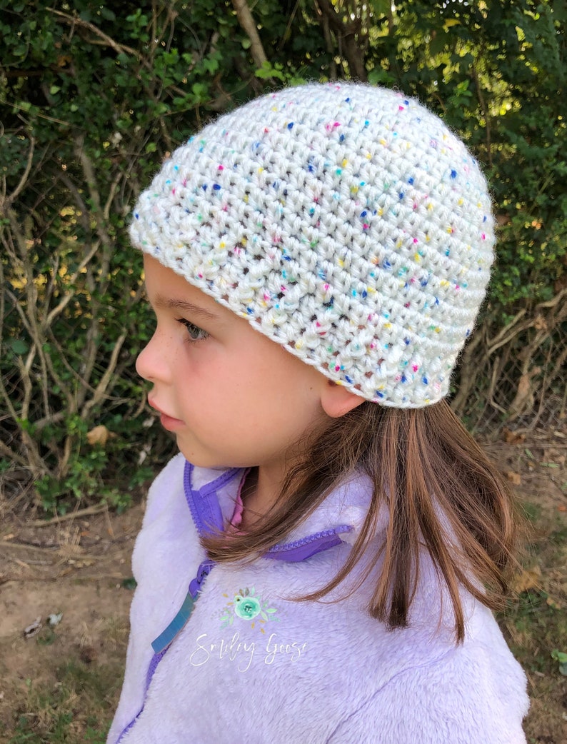 May include: A white crochet hat with colorful flecks. The hat is worn by a child with long brown hair. The child is wearing a purple fleece jacket with the text "Smiling Goose" embroidered on the front.