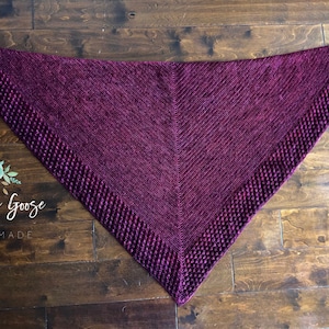 May include: A triangular purple knitted shawl with a textured border. The shawl is laid flat on a wooden surface.