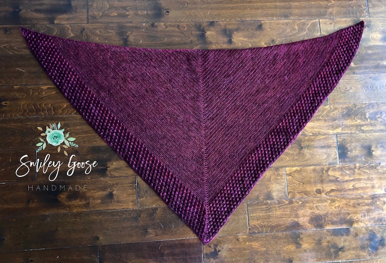 May include: A triangular purple knitted shawl with a textured border. The shawl is laid flat on a wooden surface.