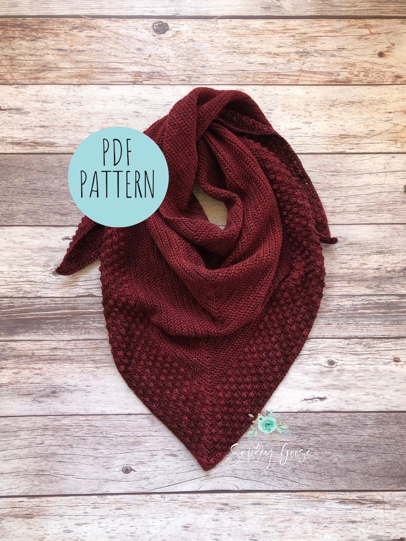 May include: A burgundy crochet shawl with a textured stitch pattern. The shawl is folded and displayed on a wooden surface. A circle with the text "PDF PATTERN" is in the upper left corner of the image.