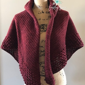 May include: A maroon crocheted shawl with a hood and a textured border. The shawl is draped over a mannequin.