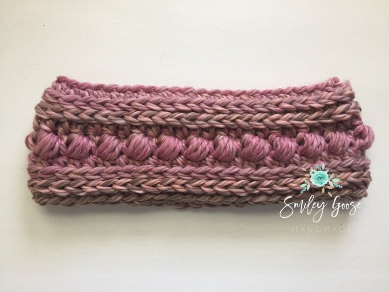 Puede incluir: Una diadema de crochet rosa con un dise&ntilde;o texturizado. La diadema tiene una fila de peque&ntilde;os bultos en relieve.