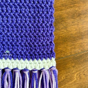 Puede incluir: Un tejido de ganchillo morado con un patr&oacute;n de puntos texturizados, una banda horizontal de hilo color crema y un fleco compuesto por hebras alternas de hilo morado oscuro, morado claro y blanco. El texto Smiley Goose es visible.