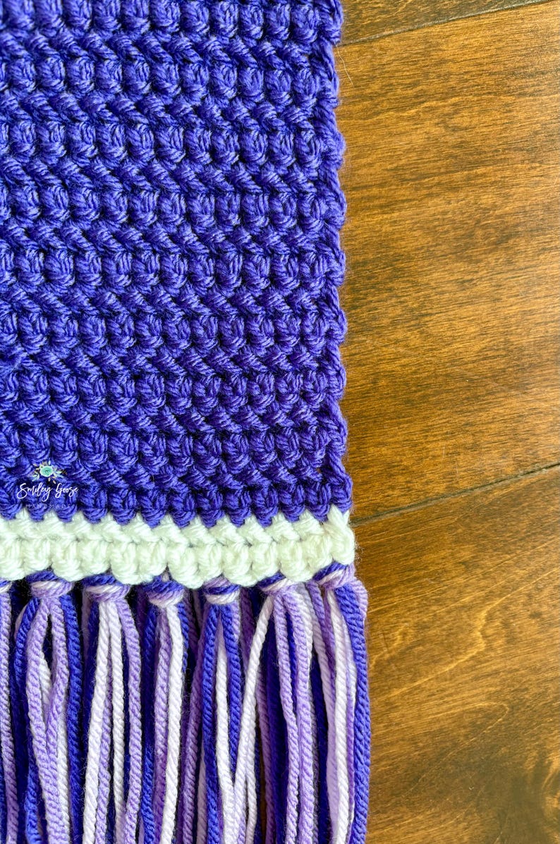 Puede incluir: Un tejido de ganchillo morado con un patr&oacute;n de puntos texturizados, una banda horizontal de hilo color crema y un fleco compuesto por hebras alternas de hilo morado oscuro, morado claro y blanco. El texto Smiley Goose es visible.