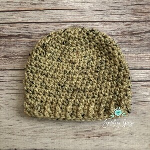 May include: A crocheted beanie hat in a green and black speckled pattern. The hat is on a wooden surface. The text "Smiley Goose" is visible in the bottom right corner of the image.