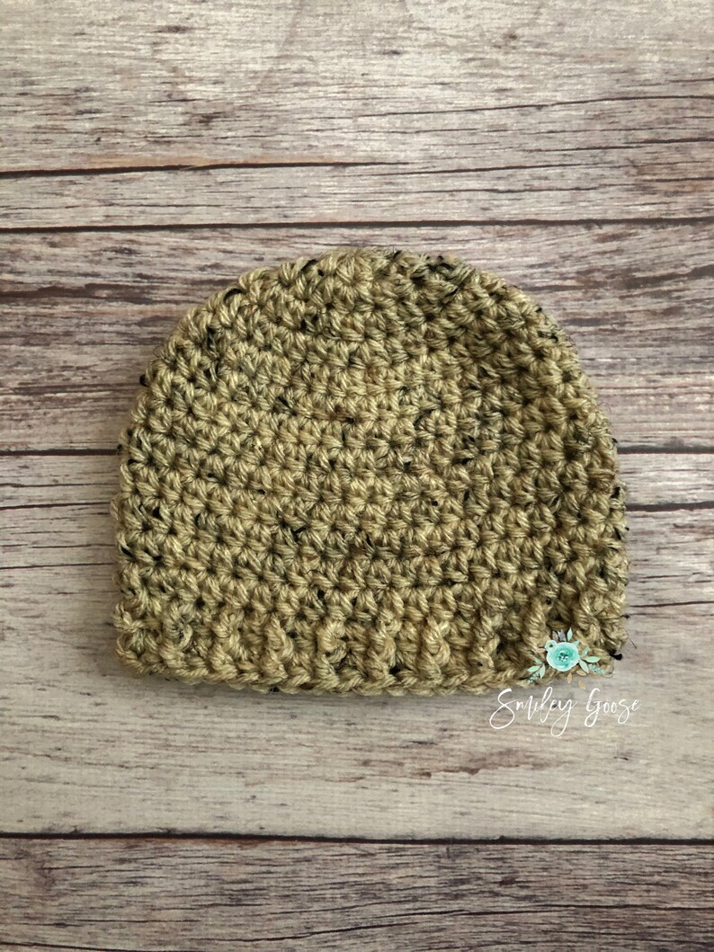 May include: A crocheted beanie hat in a green and black speckled pattern. The hat is on a wooden surface. The text "Smiley Goose" is visible in the bottom right corner of the image.