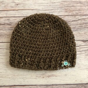May include: A brown crocheted beanie hat with a speckled texture. The hat is on a light wood background. The text "Smiley Goose" is visible in the bottom right corner of the image.