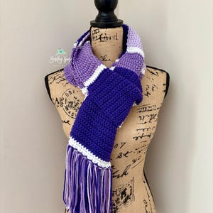Puede incluir: Una bufanda de ganchillo con rayas horizontales de bloques de color en tonos morado claro, morado medio, morado oscuro y blanco. La bufanda est&aacute; terminada con largos flecos multicolores en los extremos, mostrando una textura de hilo.