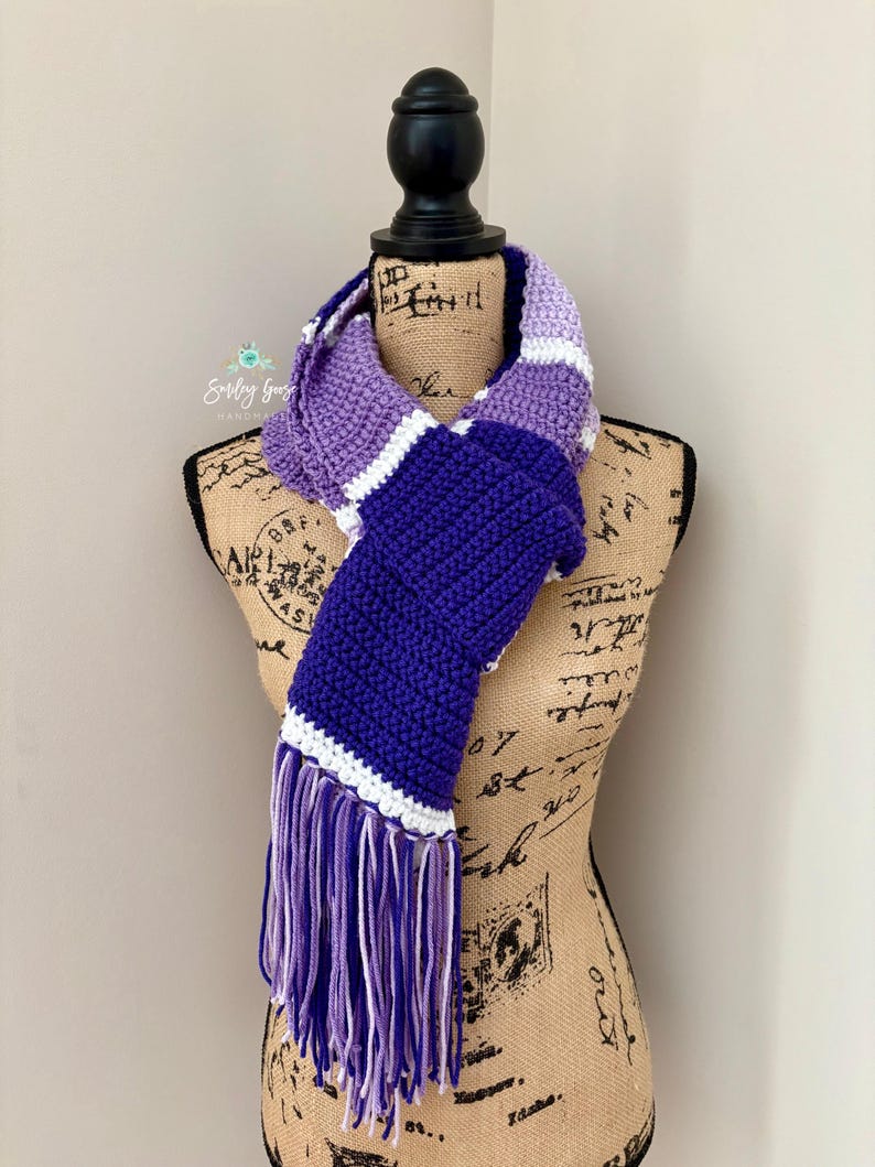 Puede incluir: Una bufanda de ganchillo con rayas horizontales de bloques de color en tonos morado claro, morado medio, morado oscuro y blanco. La bufanda est&aacute; terminada con largos flecos multicolores en los extremos, mostrando una textura de hilo.