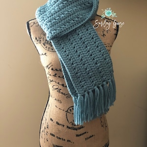 May include: A teal blue crocheted scarf with fringe on a fabric covered mannequin. The scarf is folded and draped around the mannequin's neck. The mannequin is standing on a black stand.