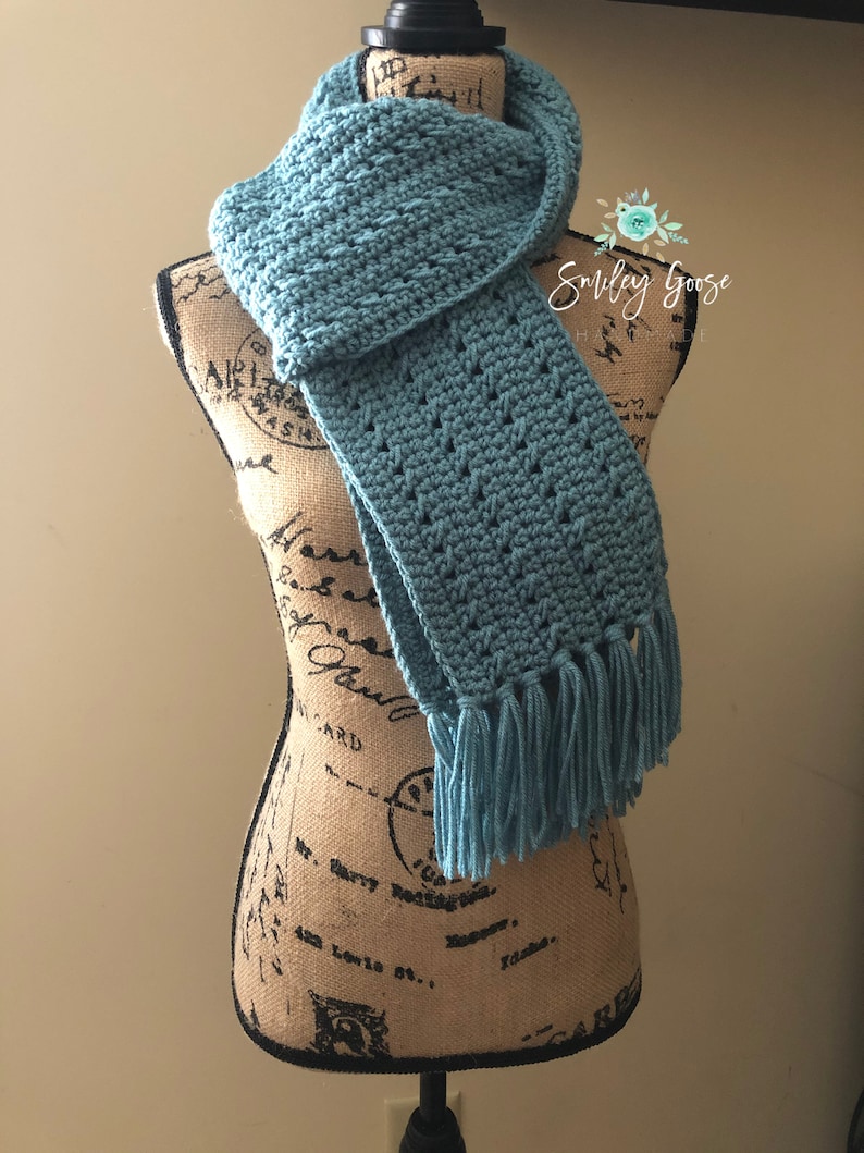 May include: A teal blue crocheted scarf with fringe on a fabric covered mannequin. The scarf is folded and draped around the mannequin's neck. The mannequin is standing on a black stand.