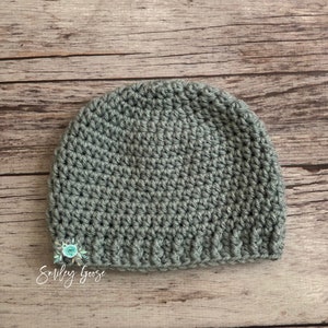 May include: A sage green crocheted beanie hat on a wood background. The hat is made with a simple stitch and has a folded brim.