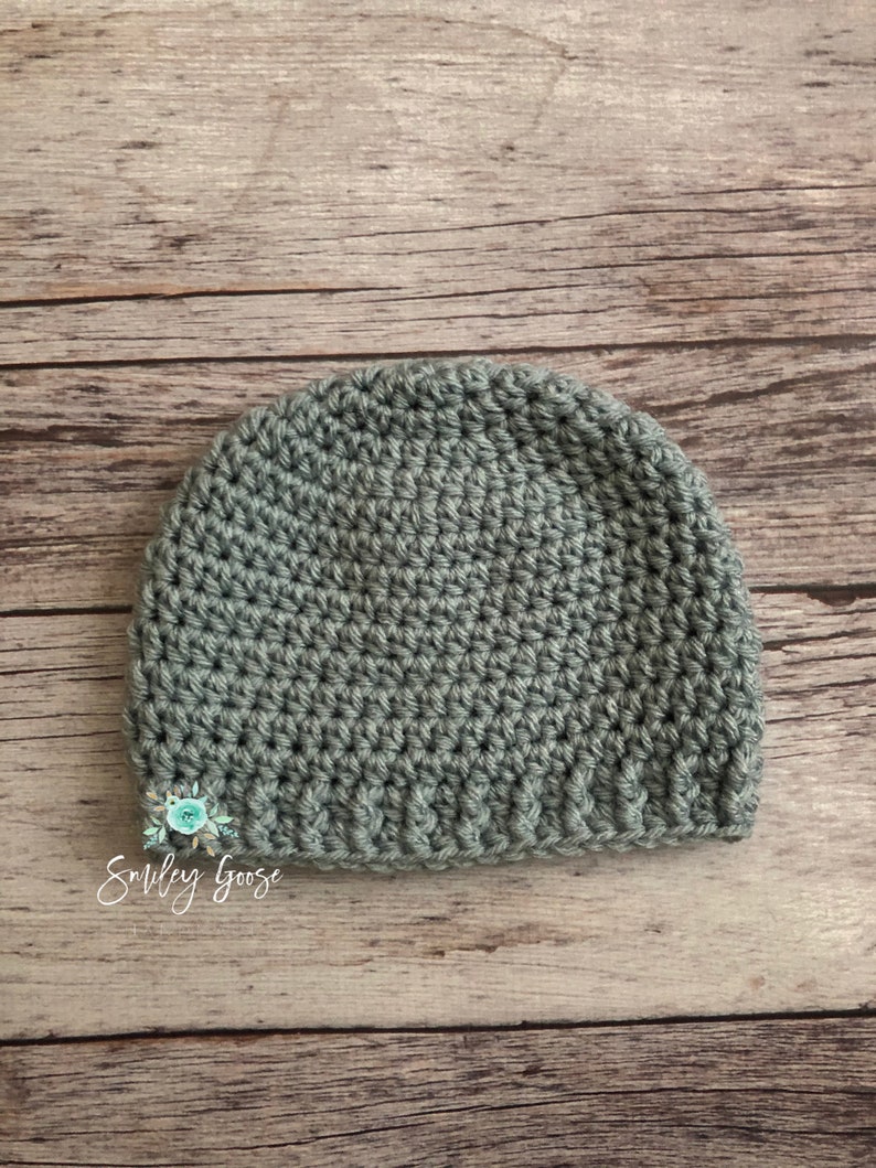 May include: A sage green crocheted beanie hat on a wood background. The hat is made with a simple stitch and has a folded brim.