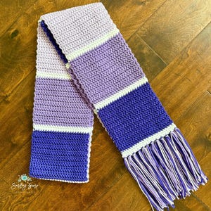 Puede incluir: Una bufanda de ganchillo larga y rectangular con rayas horizontales de bloques de color en tonos lavanda claro, morado medio y violeta intenso, separadas por finas bandas blancas. La bufanda termina con largos flecos multicolores.