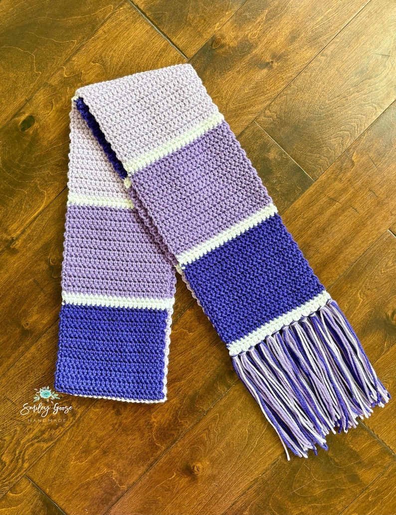 Puede incluir: Una bufanda de ganchillo larga y rectangular con rayas horizontales de bloques de color en tonos lavanda claro, morado medio y violeta intenso, separadas por finas bandas blancas. La bufanda termina con largos flecos multicolores.