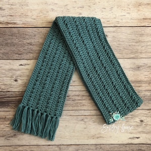 May include: A teal crocheted scarf with fringe on the ends. The scarf is laid out on a wooden surface.