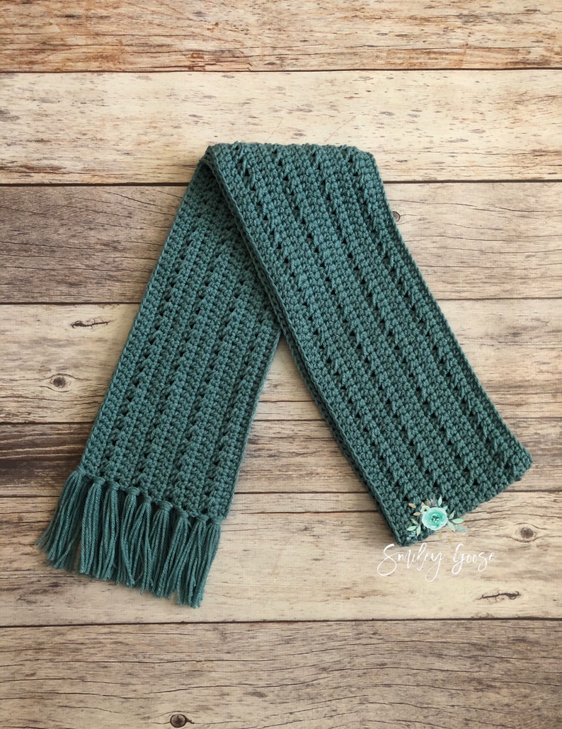 May include: A teal crocheted scarf with fringe on the ends. The scarf is laid out on a wooden surface.