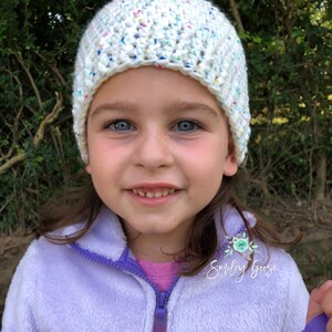 May include: A young girl wearing a white crocheted hat with colorful flecks. The hat has a folded brim. She is wearing a purple fleece jacket with a floral patch that says "Smiley Goose".