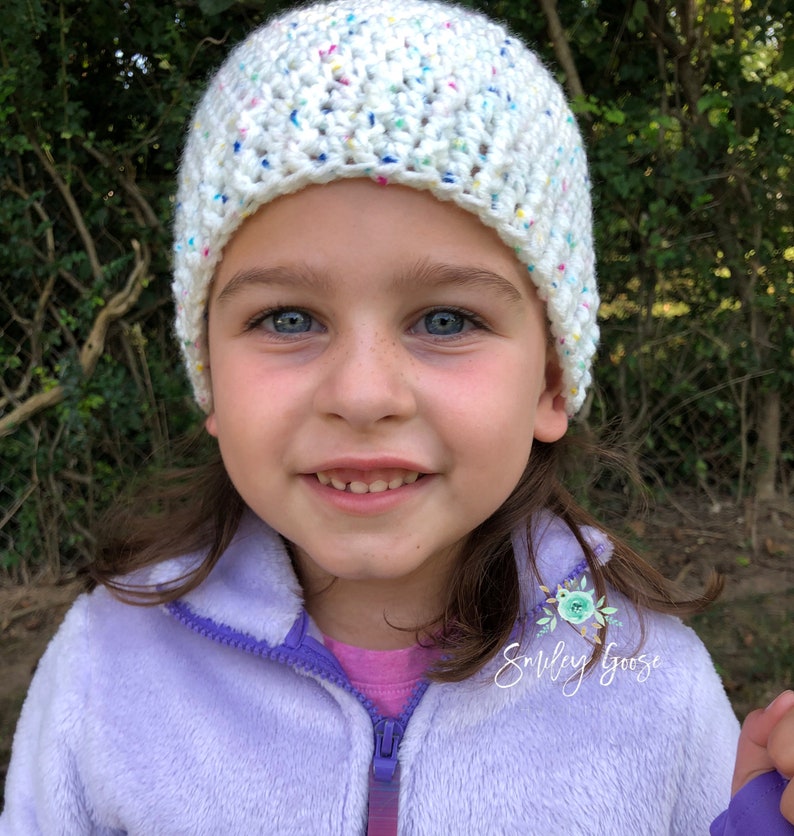 May include: A young girl wearing a white crocheted hat with colorful flecks. The hat has a folded brim. She is wearing a purple fleece jacket with a floral patch that says "Smiley Goose".