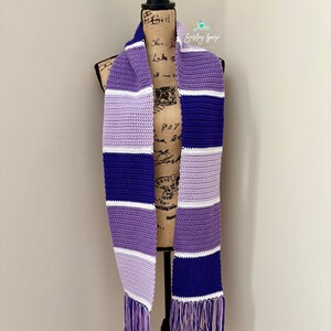 Puede incluir: Una bufanda de ganchillo con rayas horizontales en tonos morado oscuro, morado claro, lavanda y blanco. La bufanda presenta un patr&oacute;n de puntos texturizado y est&aacute; rematada con largos flecos multicolores en ambos extremos de la tela.