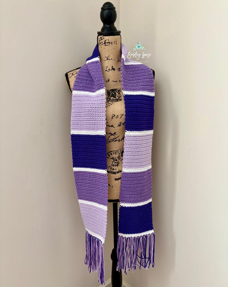 Puede incluir: Una bufanda de ganchillo con rayas horizontales en tonos morado oscuro, morado claro, lavanda y blanco. La bufanda presenta un patr&oacute;n de puntos texturizado y est&aacute; rematada con largos flecos multicolores en ambos extremos de la tela.