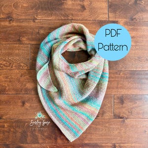 Può includere: Uno scialle triangolare lavorato a maglia con una tavolozza di colori variegati in filato verde acqua, arancione, rosa e marrone chiaro. L'articolo è mostrato con una grafica circolare azzurra contenente il testo PDF Pattern e un logo Smiley Goose HANDMADE.