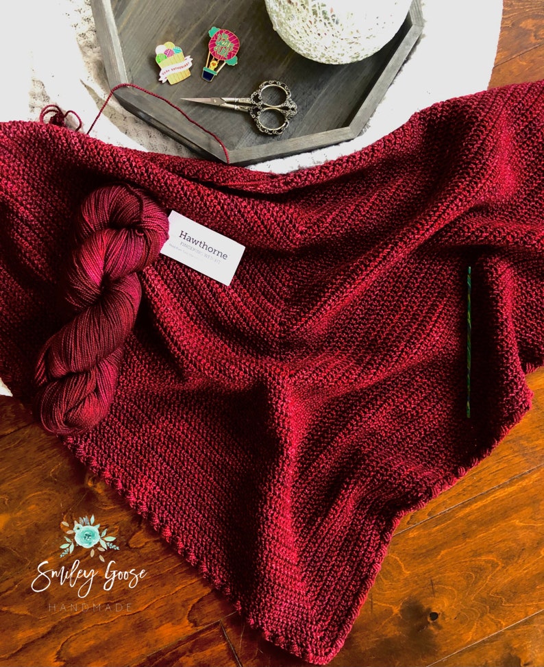 May include: A burgundy crocheted shawl with a label that reads "Hawthorne" and a tag that reads "Smiley Goose Handmade".