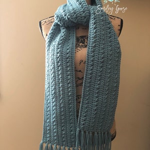 May include: A teal blue crocheted scarf with fringe on the ends. The scarf is displayed on a mannequin.