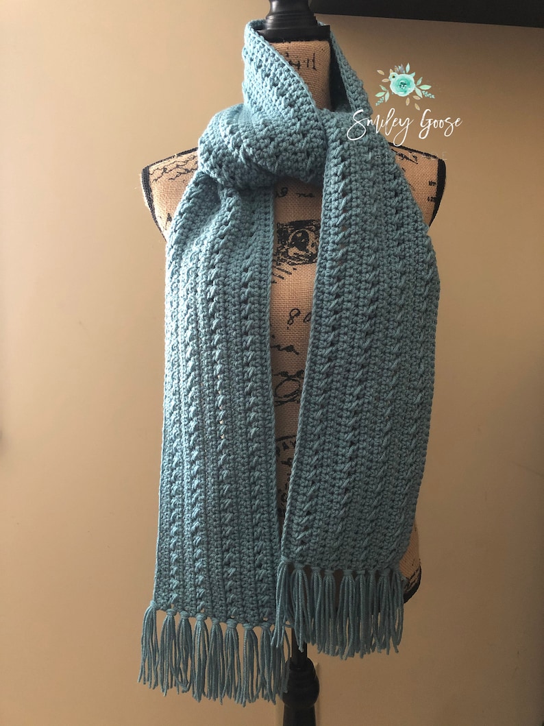 May include: A teal blue crocheted scarf with fringe on the ends. The scarf is displayed on a mannequin.