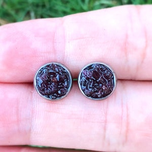 May include: A pair of silver stud earrings with a dark red, almost black, stone set in each. The stones have a rough, textured surface.