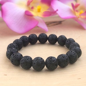 May include: A black lava stone bracelet with round, porous beads. The bracelet is displayed on a light-colored wooden surface, with pink and purple flowers in the background. The bracelet is a piece of jewelry.