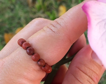 Könnte beinhalten: Nahaufnahme einer Hand, die einen roten Perlenring trägt. Der Ring besteht aus kleinen, runden Perlen, die auf einer dünnen Schnur aufgereiht sind. Der Ring wird am Zeigefinger getragen.