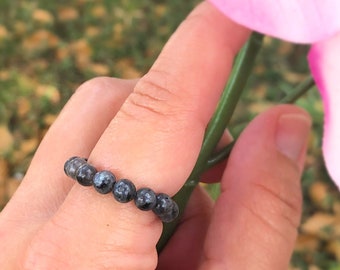Peut inclure: Une bague en perles noires avec un motif moucheté, portée sur un doigt tenant une fleur rose.