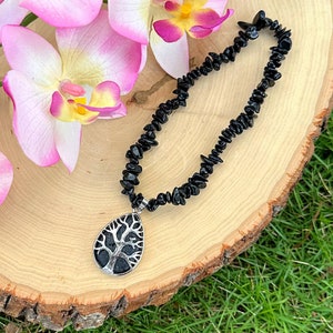 May include: Black gemstone necklace with a silver tree of life pendant. The necklace is on a wooden surface with pink orchid flowers.