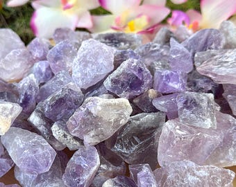 May include: A pile of raw amethyst crystals in shades of purple and lavender sits on a wooden slice. The crystals vary in size and shape, with a rough, natural texture. Pink and white flowers are in the background.