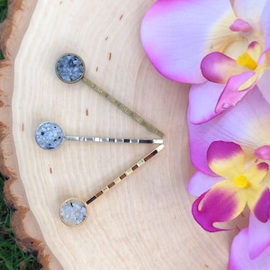 May include: Three gold hair clips with round, sparkly, stone-like embellishments. The clips are arranged in a triangle shape on a wooden surface with pink orchid flowers.