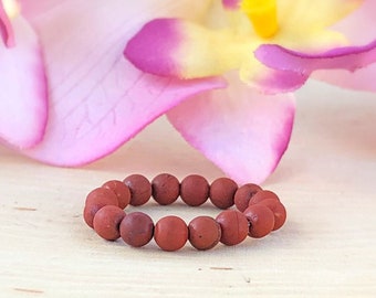 Könnte beinhalten: Ein rotes Perlenarmband aus kleinen, runden Perlen. Das Armband liegt auf einer Holzoberfläche mit rosa Blumen im Hintergrund.