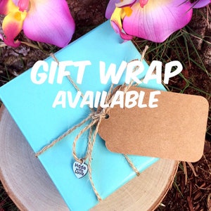 May include: A light blue gift box wrapped with twine and a brown tag. The text "GIFT WRAP AVAILABLE" is displayed on the box. A small silver heart charm with the words "Made with Love" is attached to the twine.