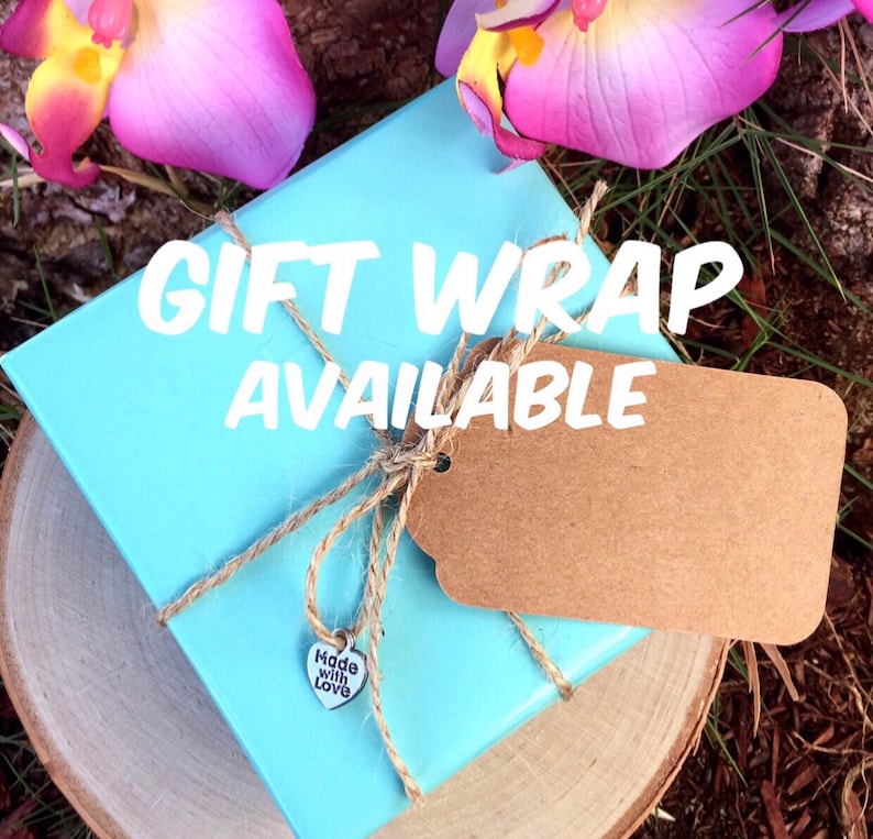 May include: A light blue gift box wrapped with twine and a brown tag. The text "GIFT WRAP AVAILABLE" is displayed on the box. A small silver heart charm with the words "Made with Love" is attached to the twine.