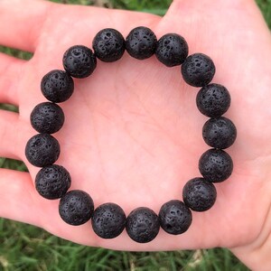May include: A black lava stone bracelet featuring round, porous beads. The bracelet is made of dark, textured volcanic rock, strung together to form a circular accessory. The beads are approximately 0.75 inches in diameter.