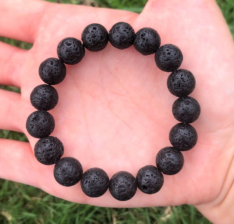 May include: A black lava stone bracelet featuring round, porous beads. The bracelet is made of dark, textured volcanic rock, strung together to form a circular accessory. The beads are approximately 0.75 inches in diameter.
