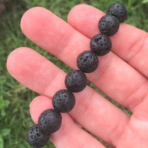 May include: A black lava stone bead bracelet. The bracelet is composed of round, porous beads. The beads are strung together, creating a simple yet striking accessory. The bracelet is held in a hand against a backdrop of green grass.