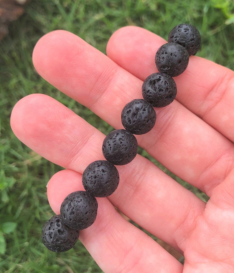 May include: A black lava stone bead bracelet. The bracelet is composed of round, porous beads. The beads are strung together, creating a simple yet striking accessory. The bracelet is held in a hand against a backdrop of green grass.
