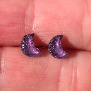 May include: A pair of crescent moon stud earrings. The earrings are a deep purple colour with a textured surface. The earrings are small and delicate, suitable for everyday wear.