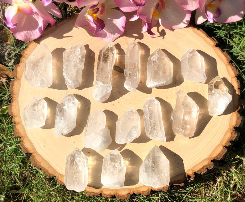 Peut inclure: Une collection de pointes de cristal de quartz clair dispos&eacute;es sur une tranche de bois ronde, avec des orchid&eacute;es roses en arri&egrave;re-plan. Les cristaux varient en taille et en forme, avec des formations naturelles et des pointes. Les cristaux sont expos&eacute;s sur une surface en bois.