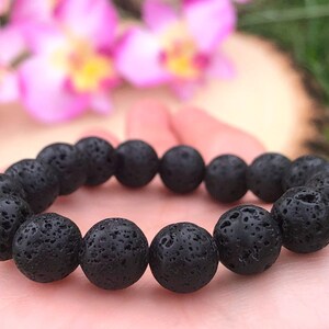 May include: A black lava stone bracelet with round, porous beads. The bracelet is held in a hand, with a blurred background of pink flowers and greenery.