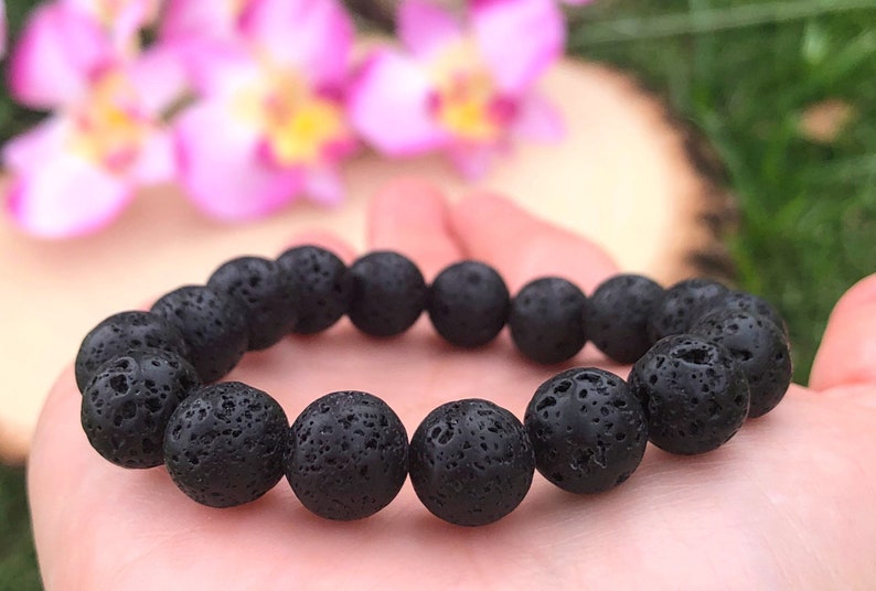 May include: A black lava stone bracelet with round, porous beads. The bracelet is held in a hand, with a blurred background of pink flowers and greenery.