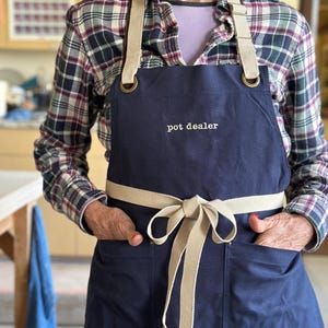 May include: A navy blue apron with a tan strap and a tan bow. The apron has two pockets and the text "pot dealer" embroidered on the front.