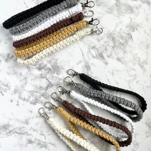 May include: A set of 10 handmade macrame keychains in various colors, including black, gray, white, brown, and yellow. Each keychain features a braided cord with a silver metal clasp.