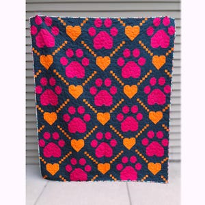 May include: A quilted blanket featuring a repeating pattern of hot pink paw prints and orange hearts set against a dark navy blue background. The design includes small orange square accents arranged in diagonal lines between the larger motifs.